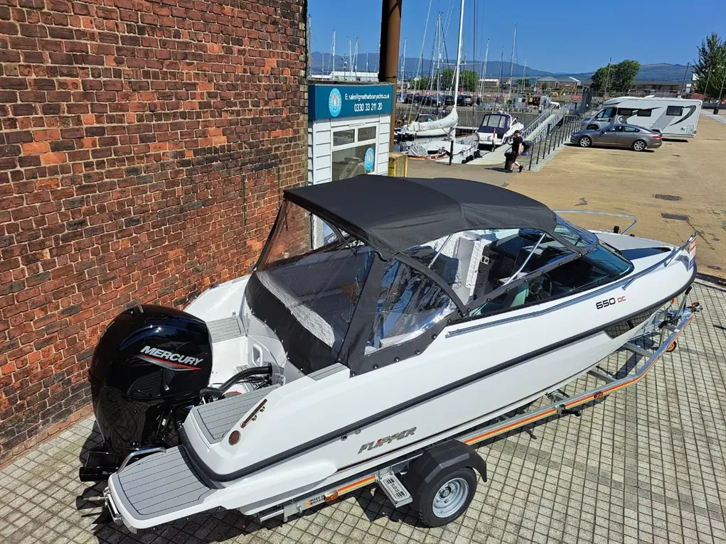 White boat on a trailer with a Mercury engine, parked near a marina with sailboats and a brick wall in the background.